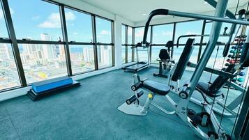 Un gimnasio con equipo de ejercicio, una banca y vistas a un paisaje urbano a través de amplias ventanas.
