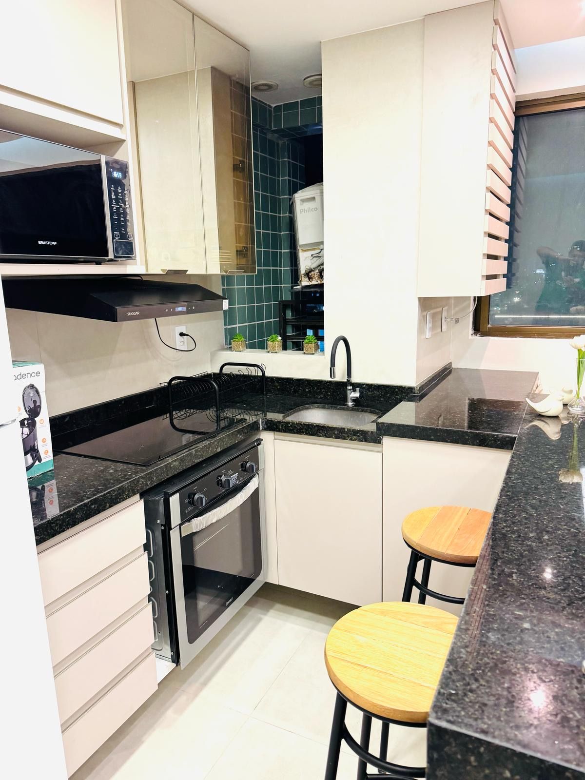 Cocina compacta con microondas, estufa y lavamanos. Hay dos taburetes y una ventana con persianas.