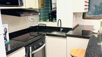 Cocina compacta con microondas, estufa y lavamanos. Hay dos taburetes y una ventana con persianas.