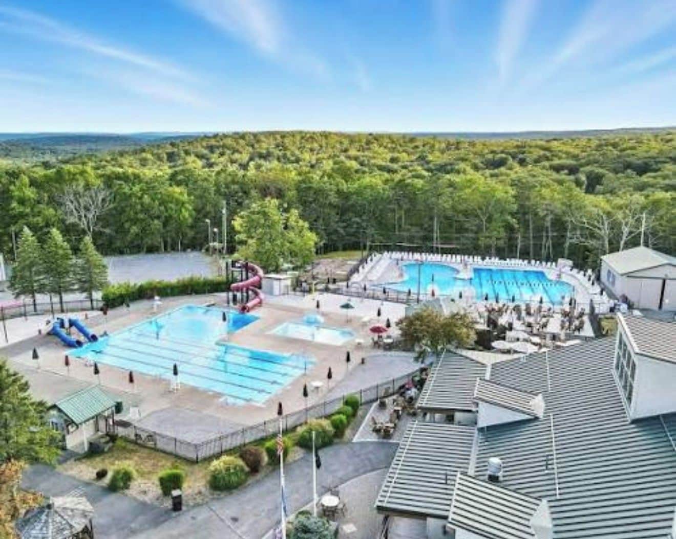 Ein Poolkomplex mit mehreren Becken und einer Wasserrutsche, umgeben von Bäumen und blauem Himmel.