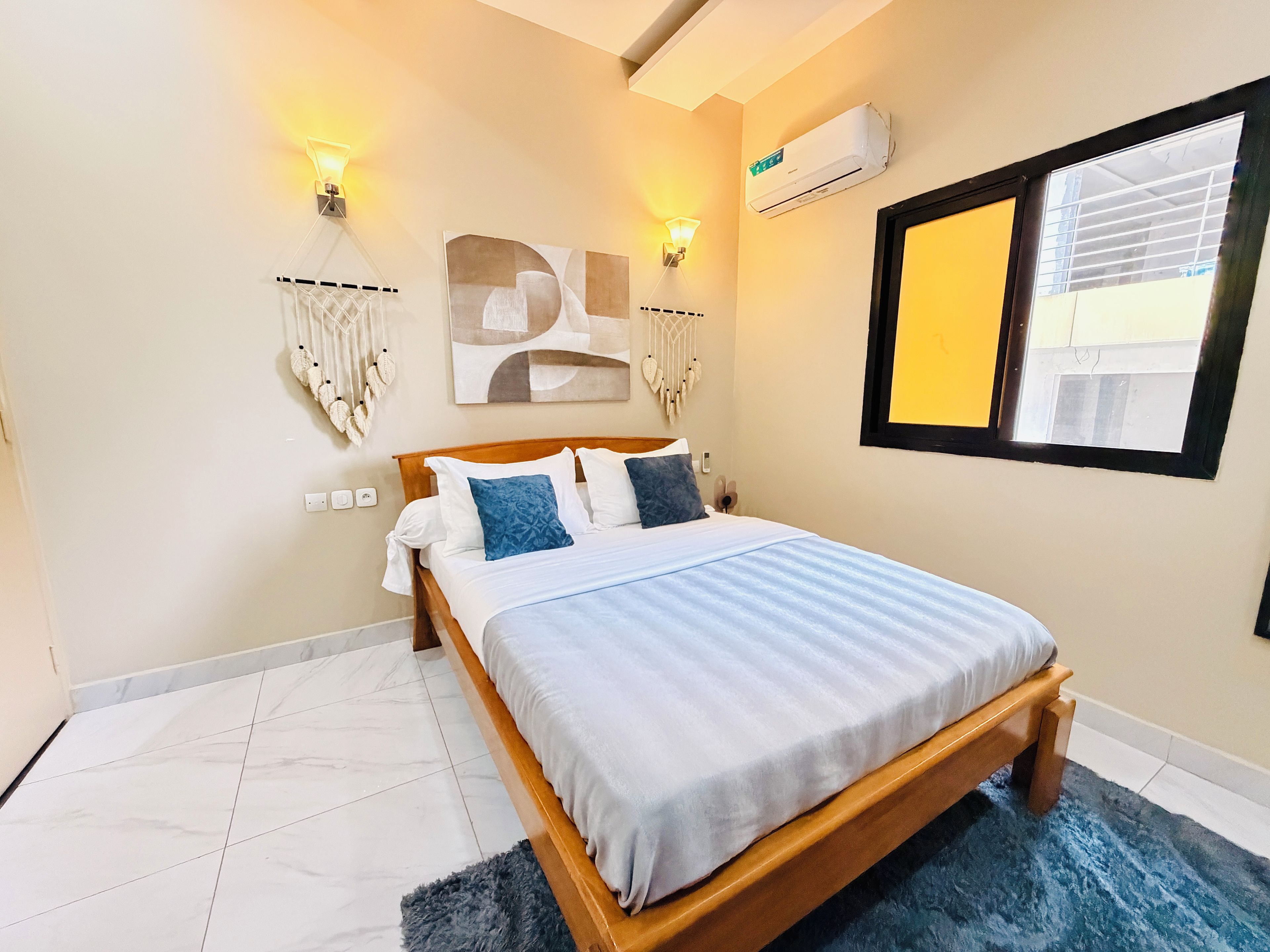 A bedroom with a wooden bed, white bedding, a blue rug, wall-mounted air conditioning, and two wall sconces.