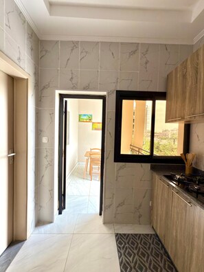 A kitchen with marble walls, a window with a view, and a dining area visible through an open door. - "Comfort & Elegance" 2-Bedroom Apartment in Bonapriso (Douala)