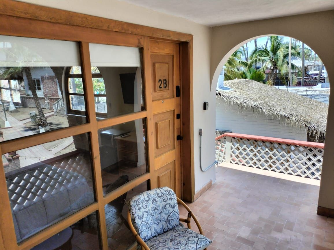 Una habitación con puerta de madera marcada '2B', ventana, silla y vista al exterior con techo de paja y palmeras.