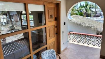 A room with a wooden door marked '2B', a window, a chair, and a view of a thatched roof and palm trees outside.