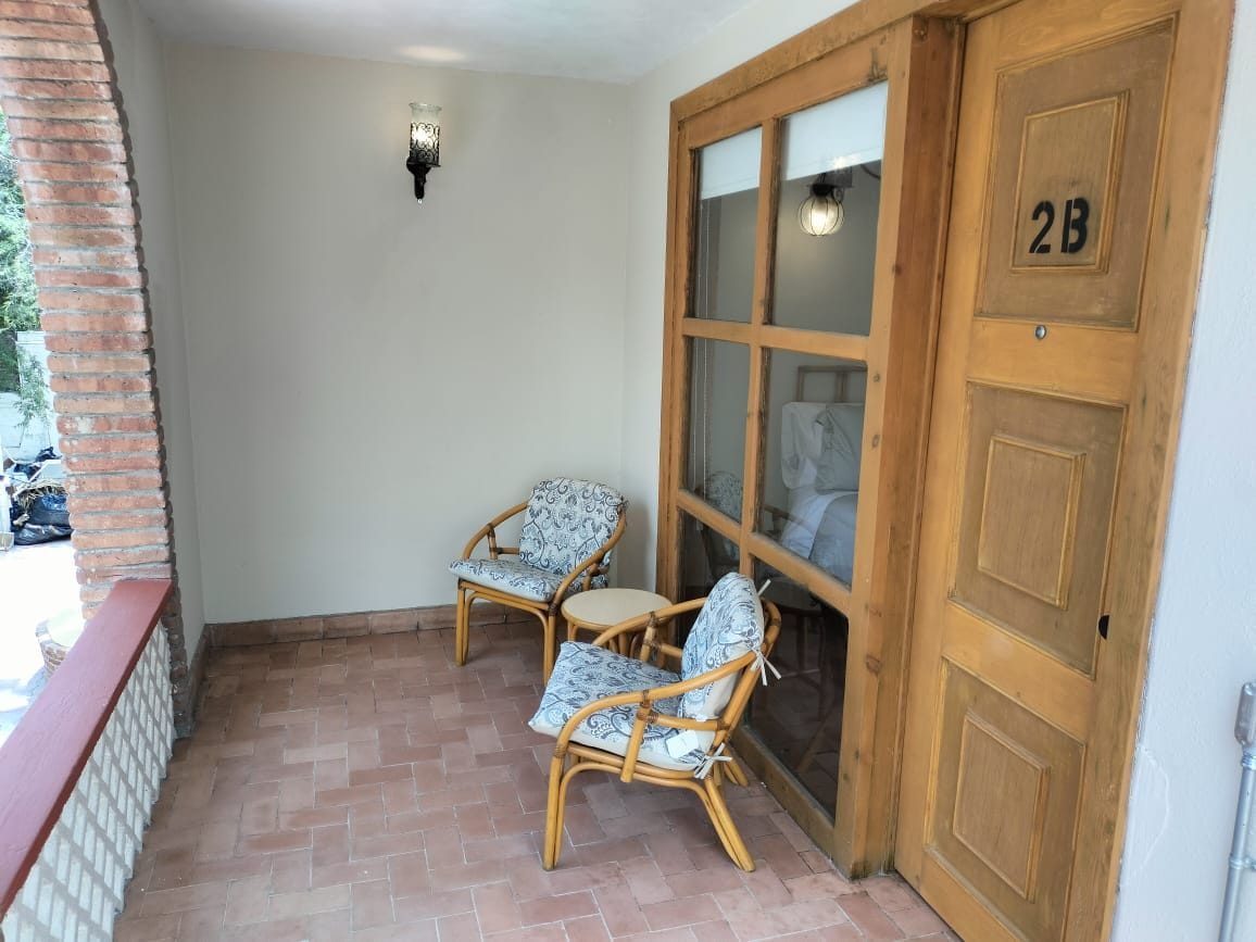 A room with a wooden door marked '2B', a glass door, two wicker chairs with patterned cushions, and a small wall-mounted light fixture.