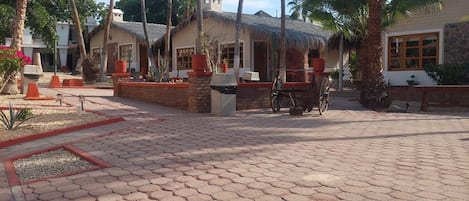 A courtyard with hexagonal paving, palm trees, and buildings with thatched roofs.