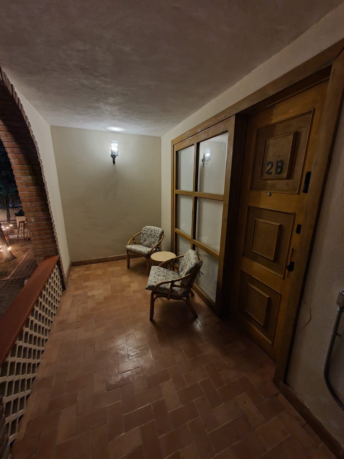 Un pasillo con una puerta de madera marcada '2B', dos sillas de mimbre, una mesita redonda y una lámpara fijada a la pared.