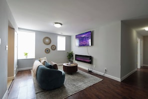 A living room with a sofa, a coffee table, a flat-screen TV mounted on the wall, and a large window. - Cozy 2BR : Forest Park • WashU • Delmar Loop (St. Louis)