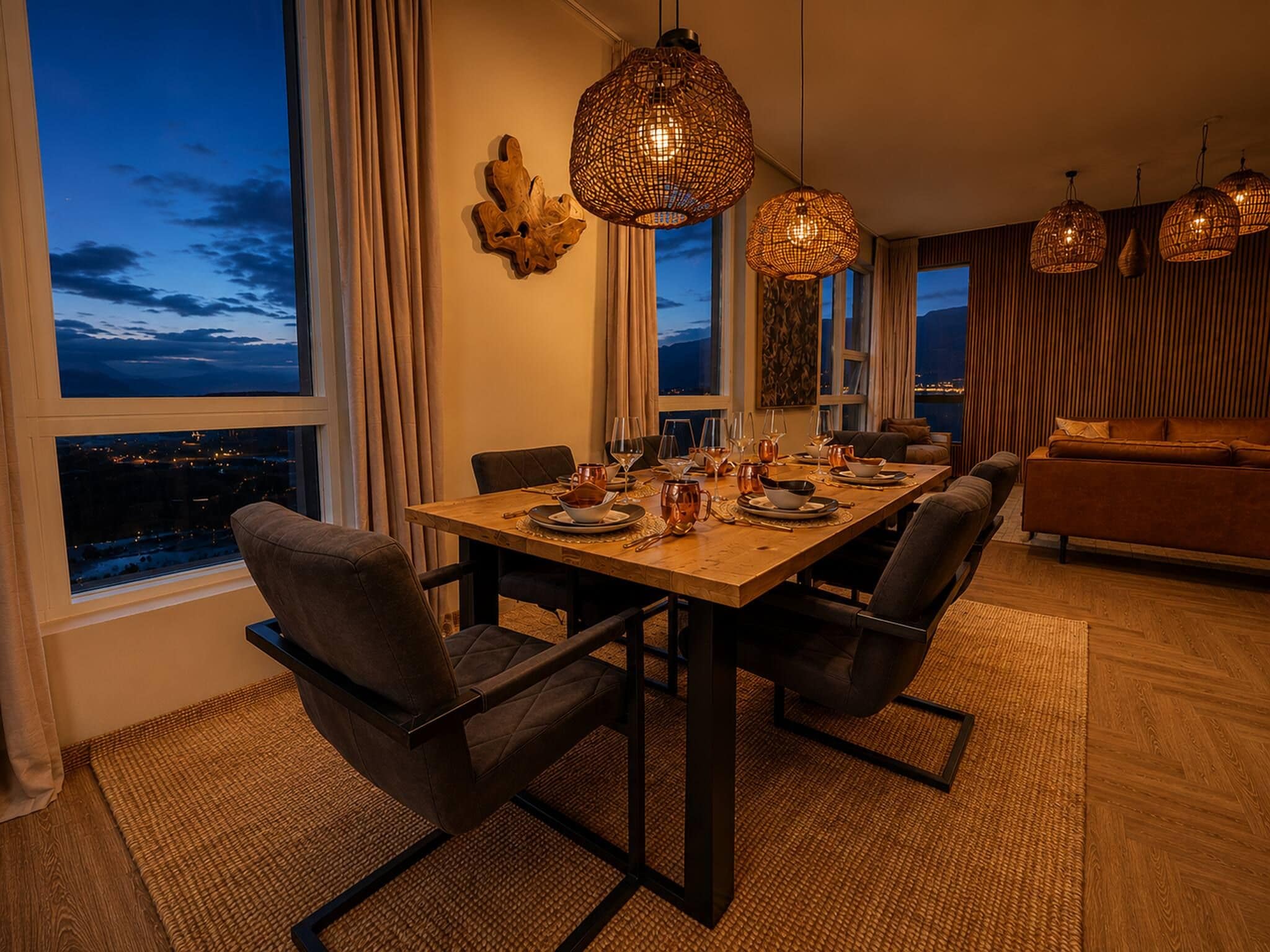Comedor con mesa de madera dispuesta para cuatro personas, sillas alrededor y vista de la ciudad a través de amplios ventanales.