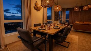 Comedor con mesa de madera dispuesta para cuatro personas, sillas alrededor y vista de la ciudad a través de amplios ventanales.