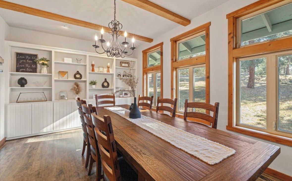A dining area with a wooden table, chairs, a chandelier, and large windows.