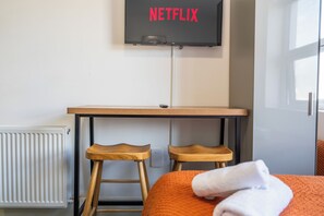 A room with a bed, a wooden desk, two wooden stools, a radiator, and a television mounted on the wall displaying 'NETFLIX'. - Sutherland Apartments - Apt 31 (London)