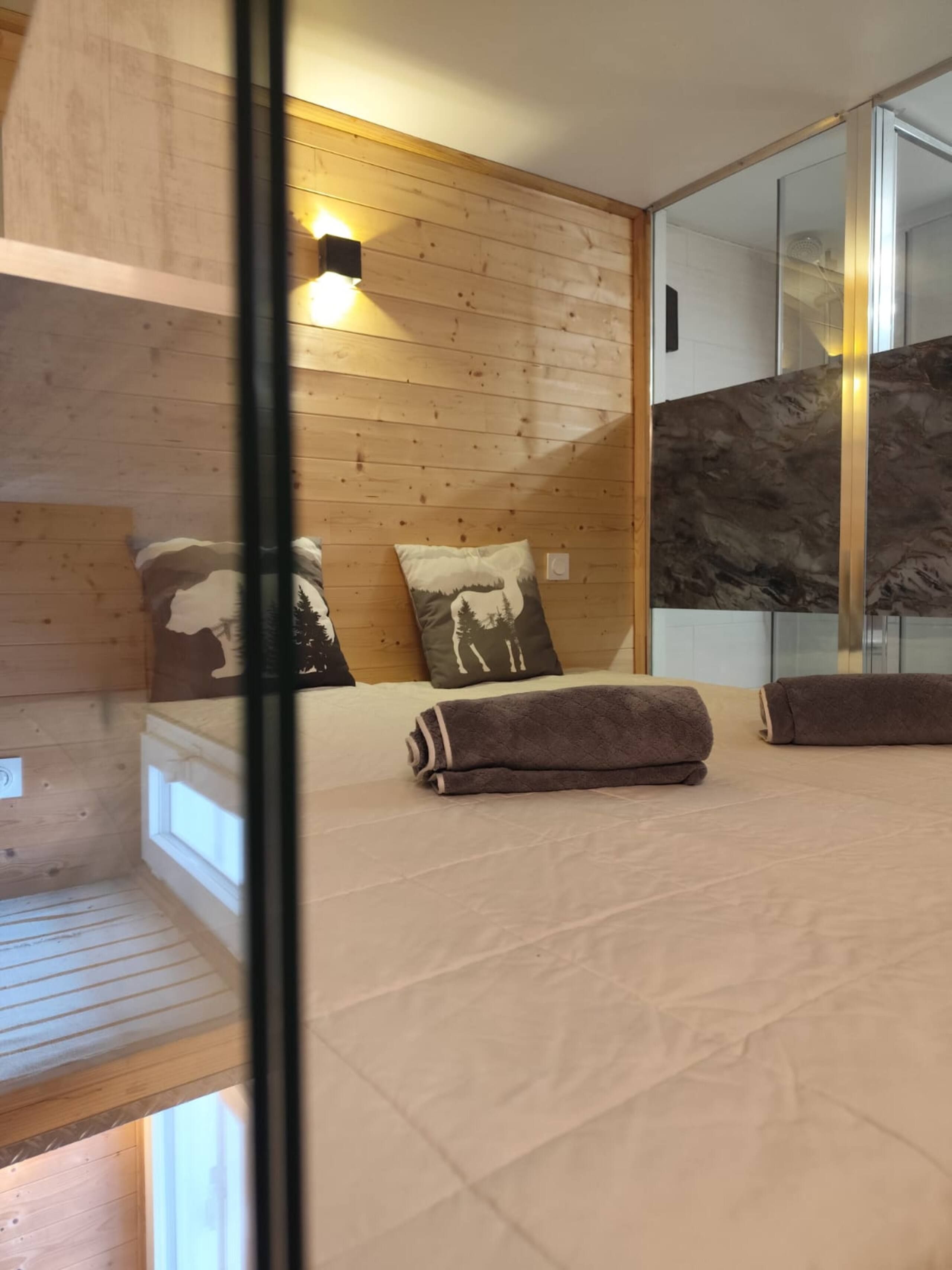 Una habitación con paredes de madera, una cama con colchón color crema, dos almohadas con diseños de montañas y un espejo grande que refleja la habitación.