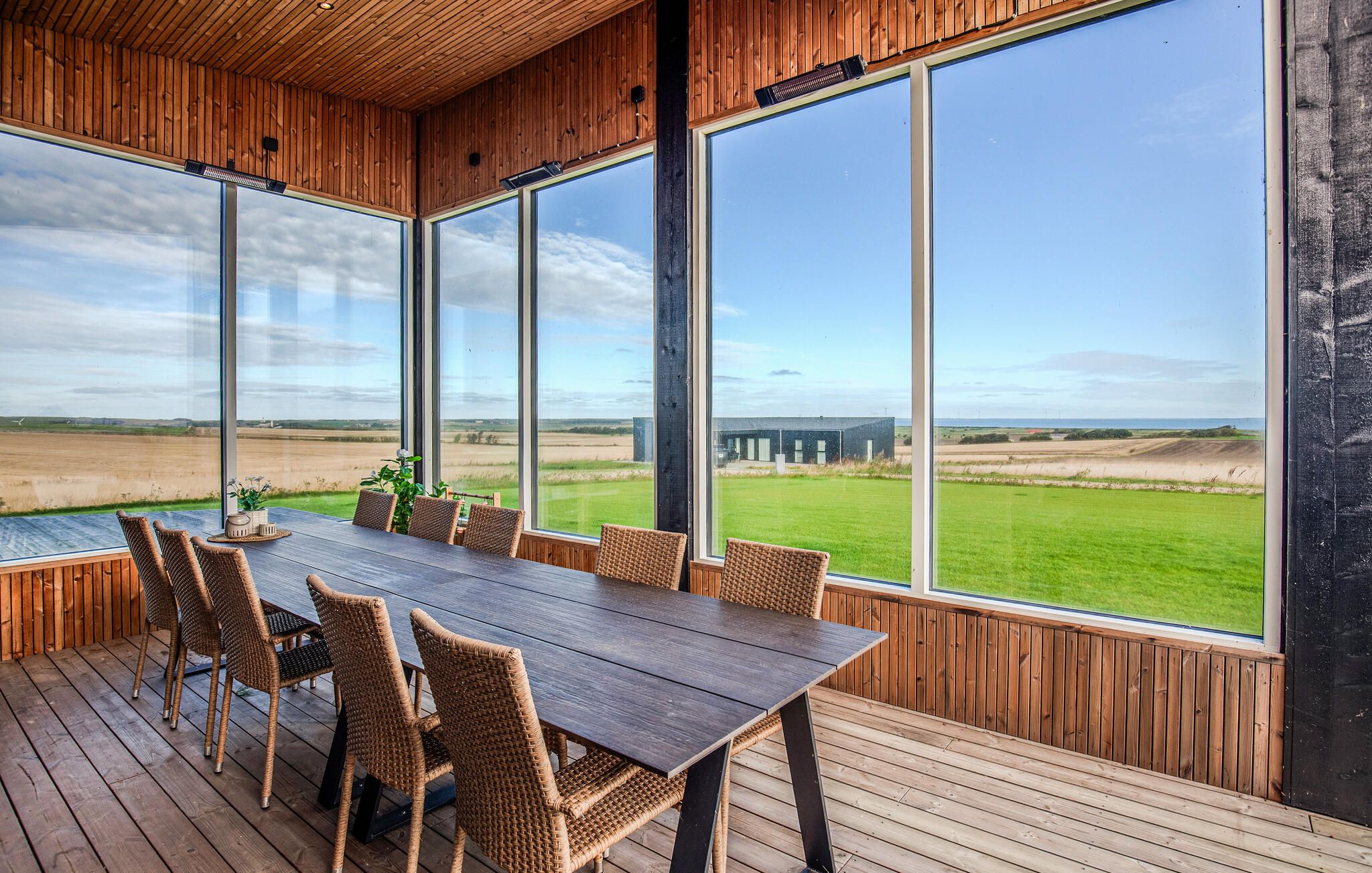En træterrasse med et bord og stole, store vinduer med udsigt over et marklandskab og fjerne bygninger.