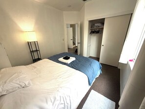 Ein Hotelzimmer mit einem Bett, einer Nachttischlampe, einem Kleiderschrank mit Kleidung und einer Tür zu einem anderen Raum.