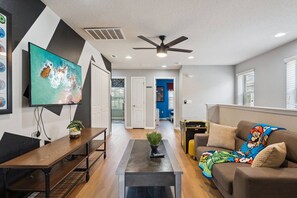 A modern living room with a flat-screen TV, a ceiling fan, and a sofa with a Mario-themed throw blanket. - Private Pool + Star Wars Game Room + Arcade Loft | Near Disney (Kissimmee)