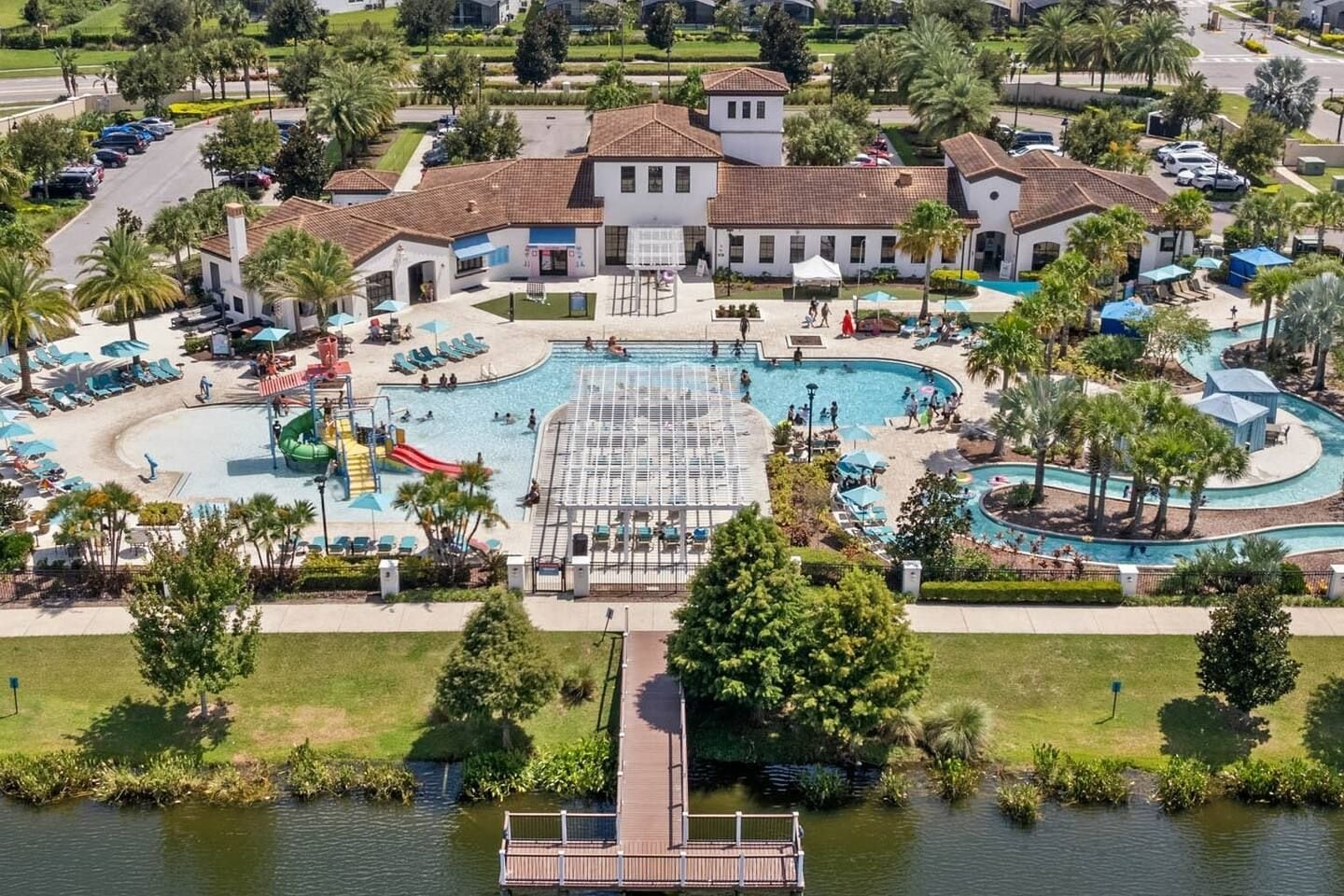 A large swimming pool with multiple lanes, surrounded by lounge chairs and umbrellas, with a bridge leading to a dock over a body of water.