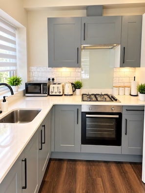 A modern kitchen with grey cabinets, a stainless steel sink, a microwave, a toaster, a kettle, and a gas stove. - Apartment Sleeps 6 With Wifi (Wembley)