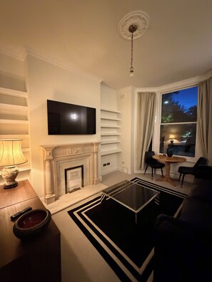 A living room with a fireplace, a television mounted on the wall, a table with chairs, and a view of the outdoors through a window. - Beautiful flat in Ealing - 4 min walk to station (London)