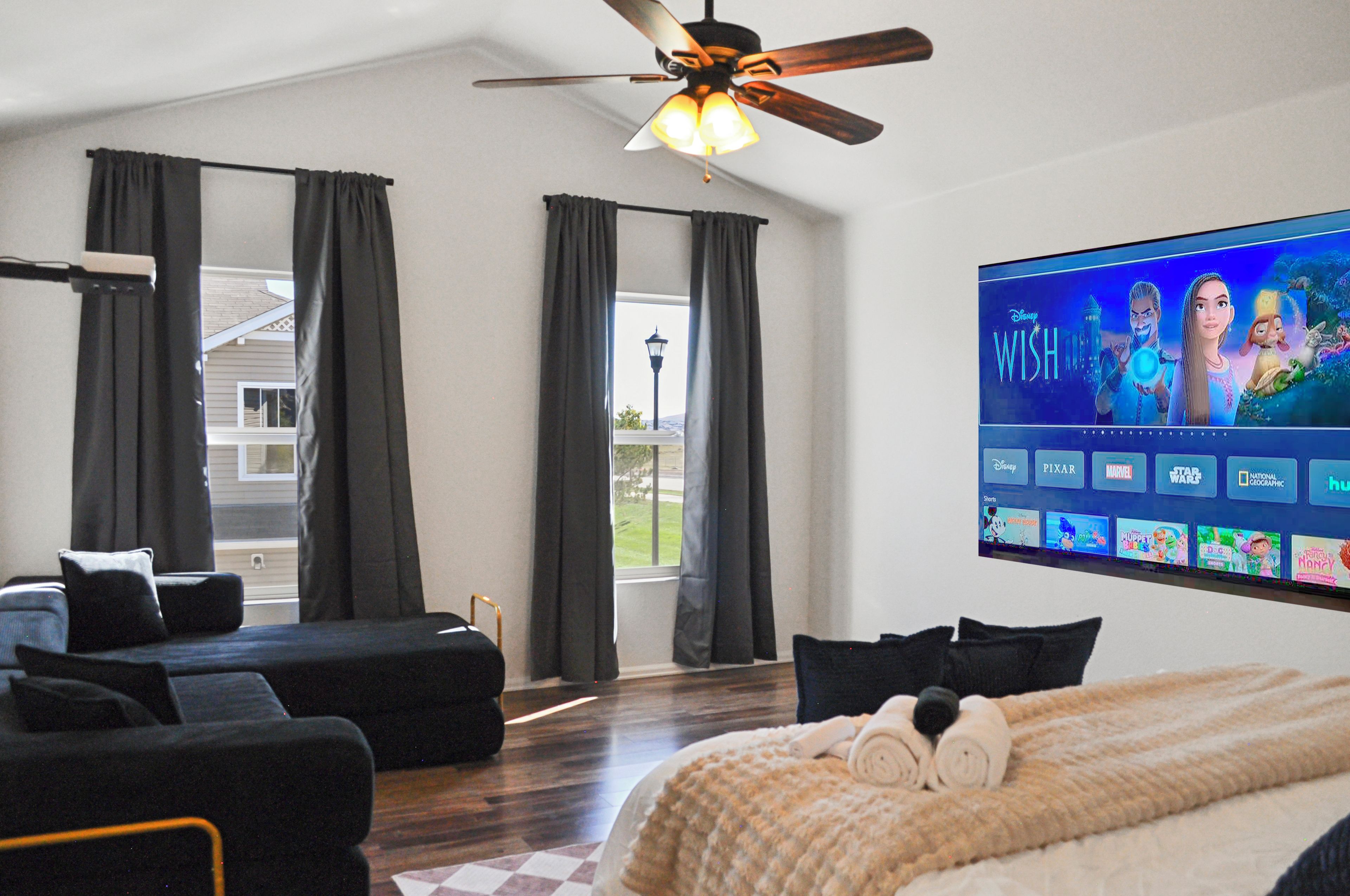 A hotel room with a large flat-screen TV displaying a movie, a ceiling fan, a sofa, a bed with a blanket, and a view of the outdoors through the windows.