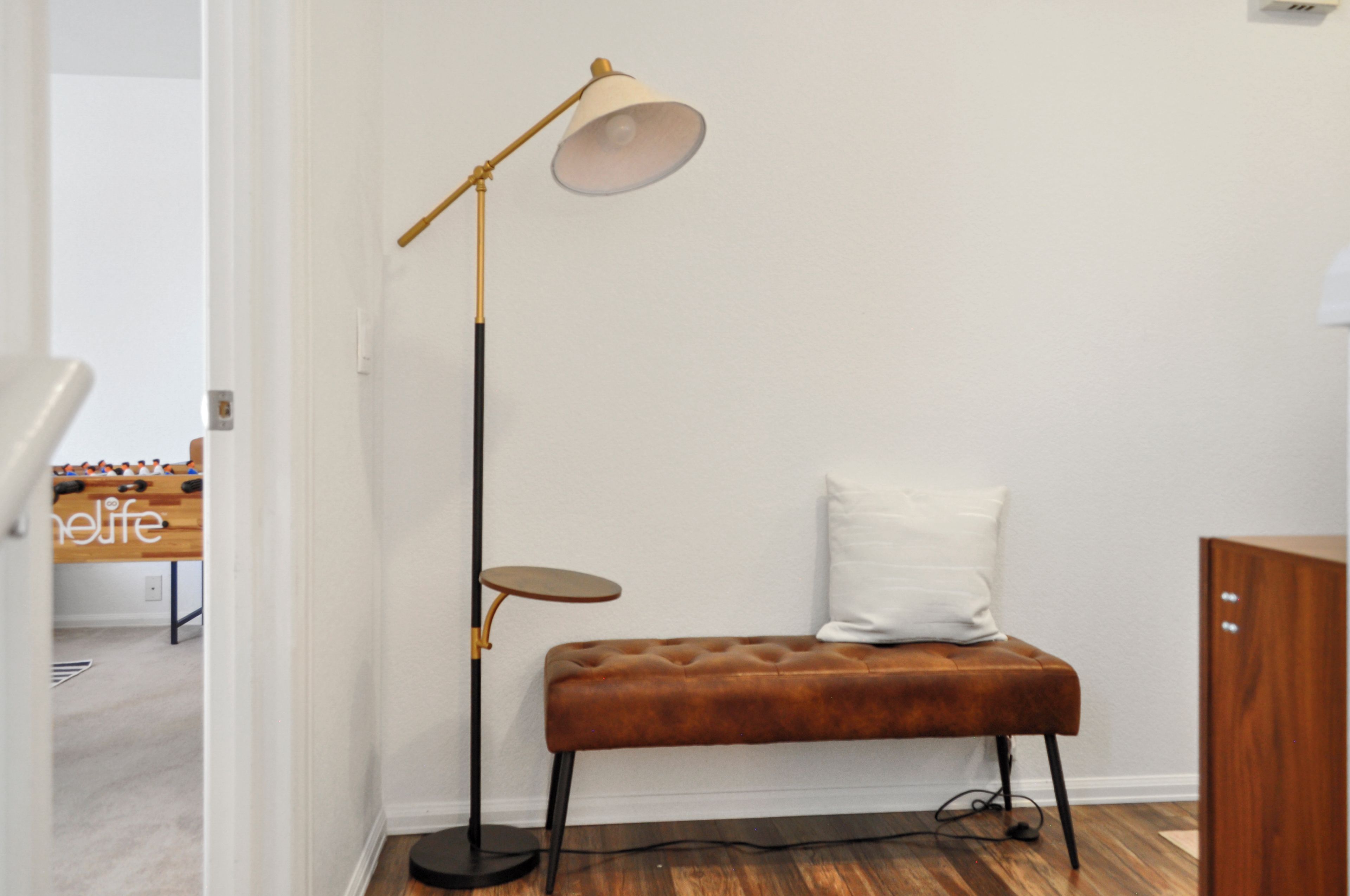 Une pièce avec un banc marron, une lampe sur pied, une petite table et un coussin blanc.