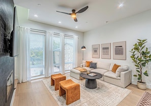 A modern living room with a sofa, coffee table, and two ottomans. There is a ceiling fan, a potted plant, and artwork on the wall. - 3BR Townhome Near Bishop Arts |Rooftop & Game Room (Dallas)