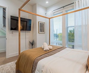 A bedroom with a four-poster bed, a flat-screen TV mounted on the wall, and a window with blinds. - 3BR Townhome Near Bishop Arts |Rooftop & Game Room (Dallas)
