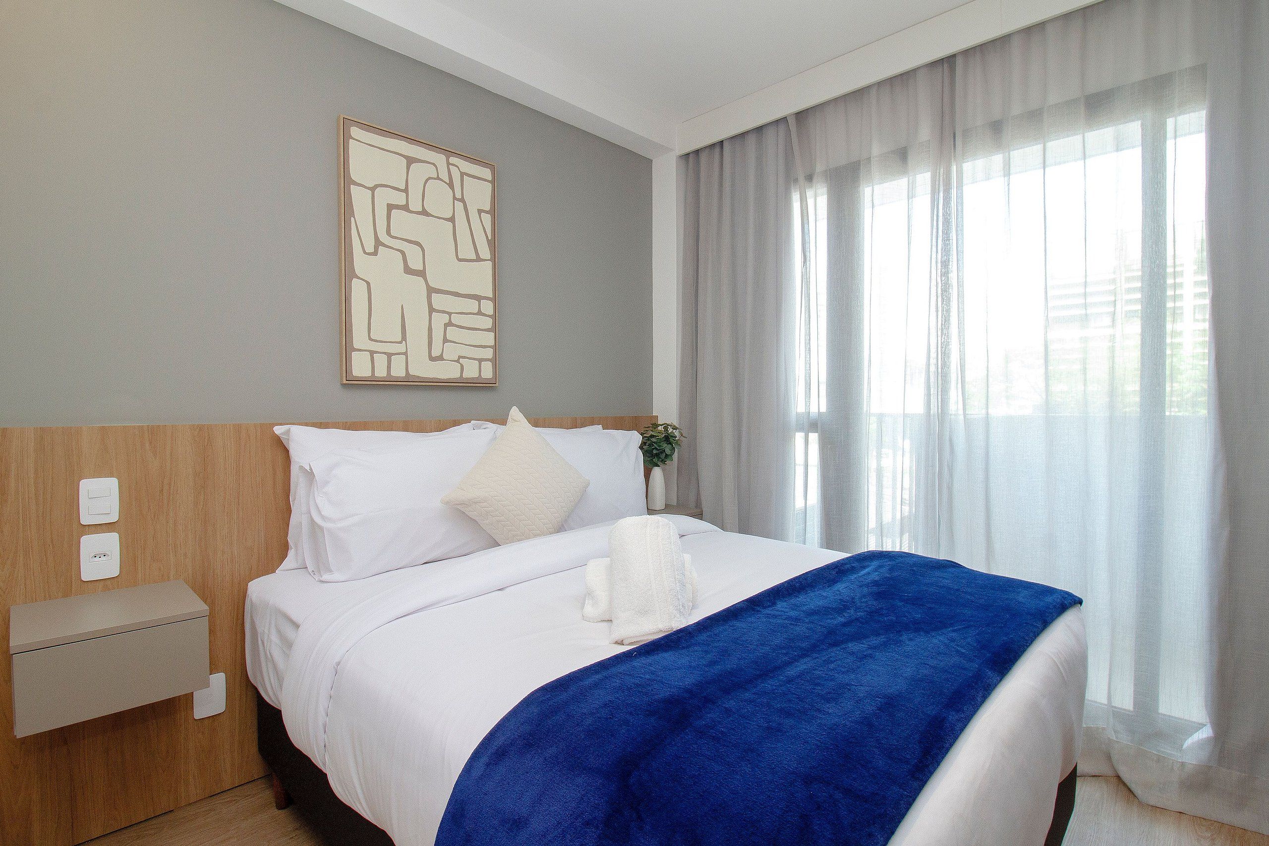 Una habitación de hotel con una cama, una mesita de noche, un enchufe y una ventana con cortinas.