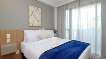 Una habitación de hotel con una cama, una mesita de noche, un enchufe y una ventana con cortinas.
