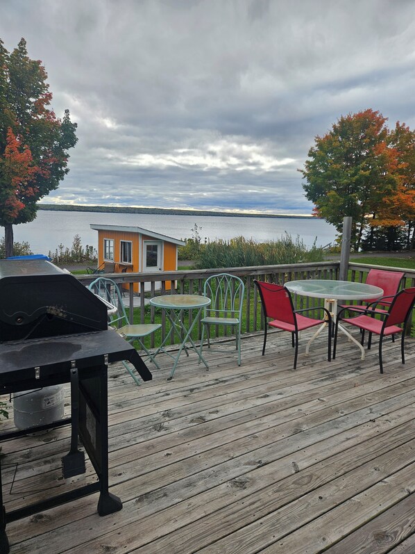 A wooden deck with outdoor furniture, a barbecue grill, and a view of a lake and a house in the background.