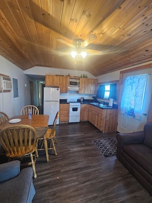 A wooden interior with a dining table, chairs, a kitchen area with cabinets and appliances, and a sofa.