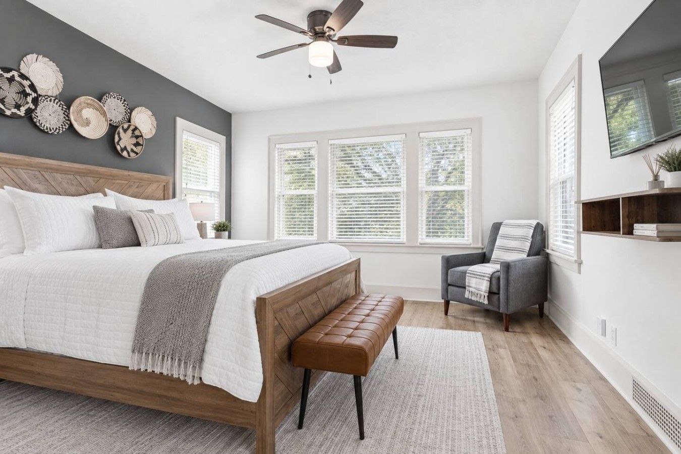 A bedroom with a bed, a bench, a chair, a ceiling fan, and a TV on the wall.