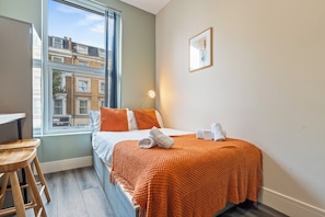 A hotel room with a bed, a desk, a window with a view of buildings, and a wall-mounted light. - Sutherland Apartments - Apt 35 (London)