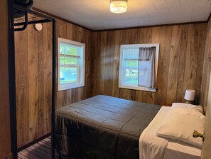 A small, single-bed room with wooden panel walls, two windows, and a bedside lamp. - Cabin 3  • 2 BR 1 BA Cabin • Sleeps 5 • Lake Roland Frontage & Trail 3 Access (Toivola)