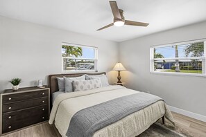 Ein Schlafzimmer mit Bett, Kommode, Nachttisch, Lampe, Deckenventilator und Fenstern mit Blick auf Palmen und ein Haus.