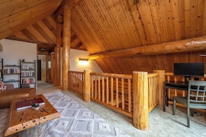 A cozy cabin interior with a wooden ceiling, a bookshelf, a fireplace, and a television.