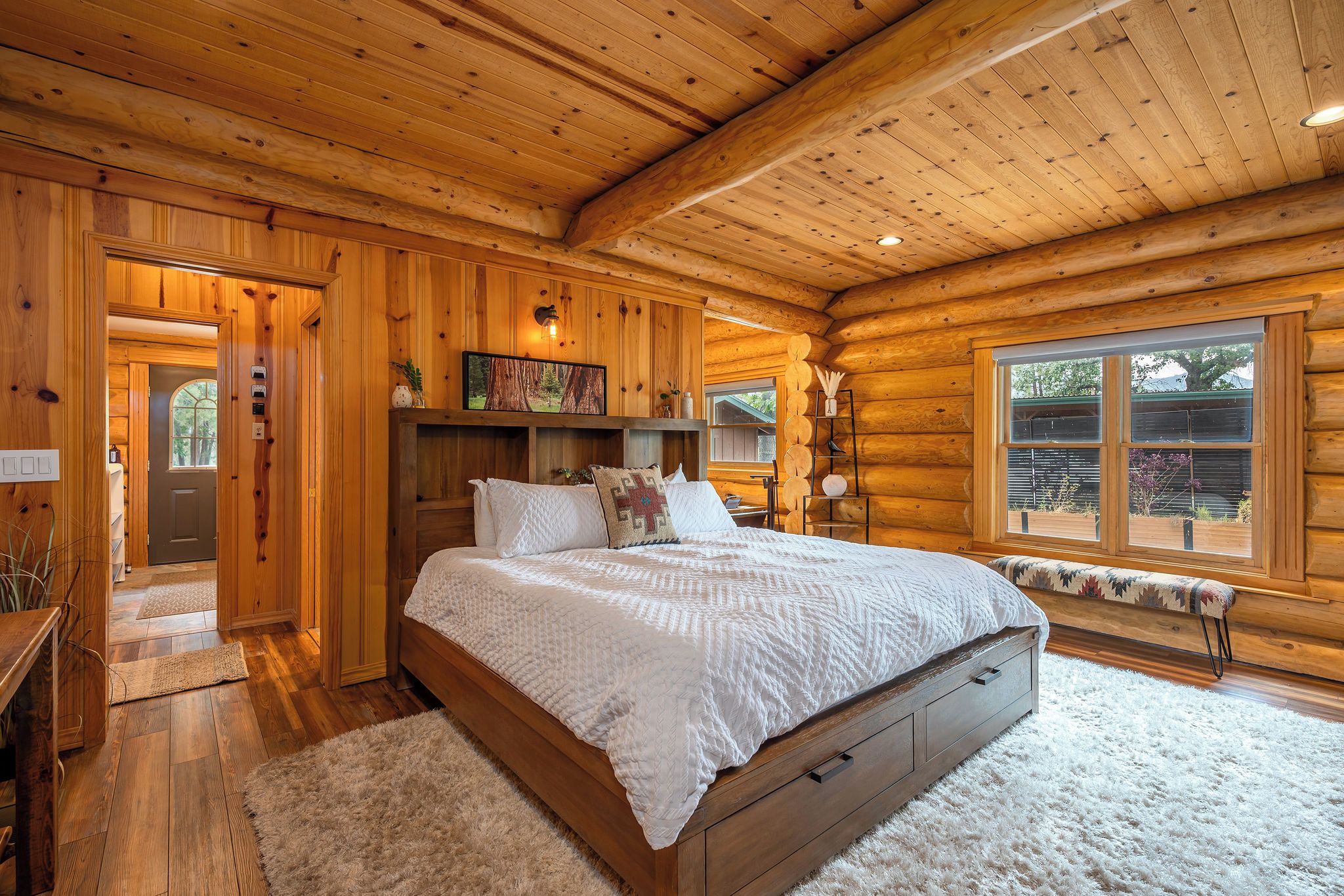 A log cabin bedroom with a large bed, wooden walls, a window, and a door.