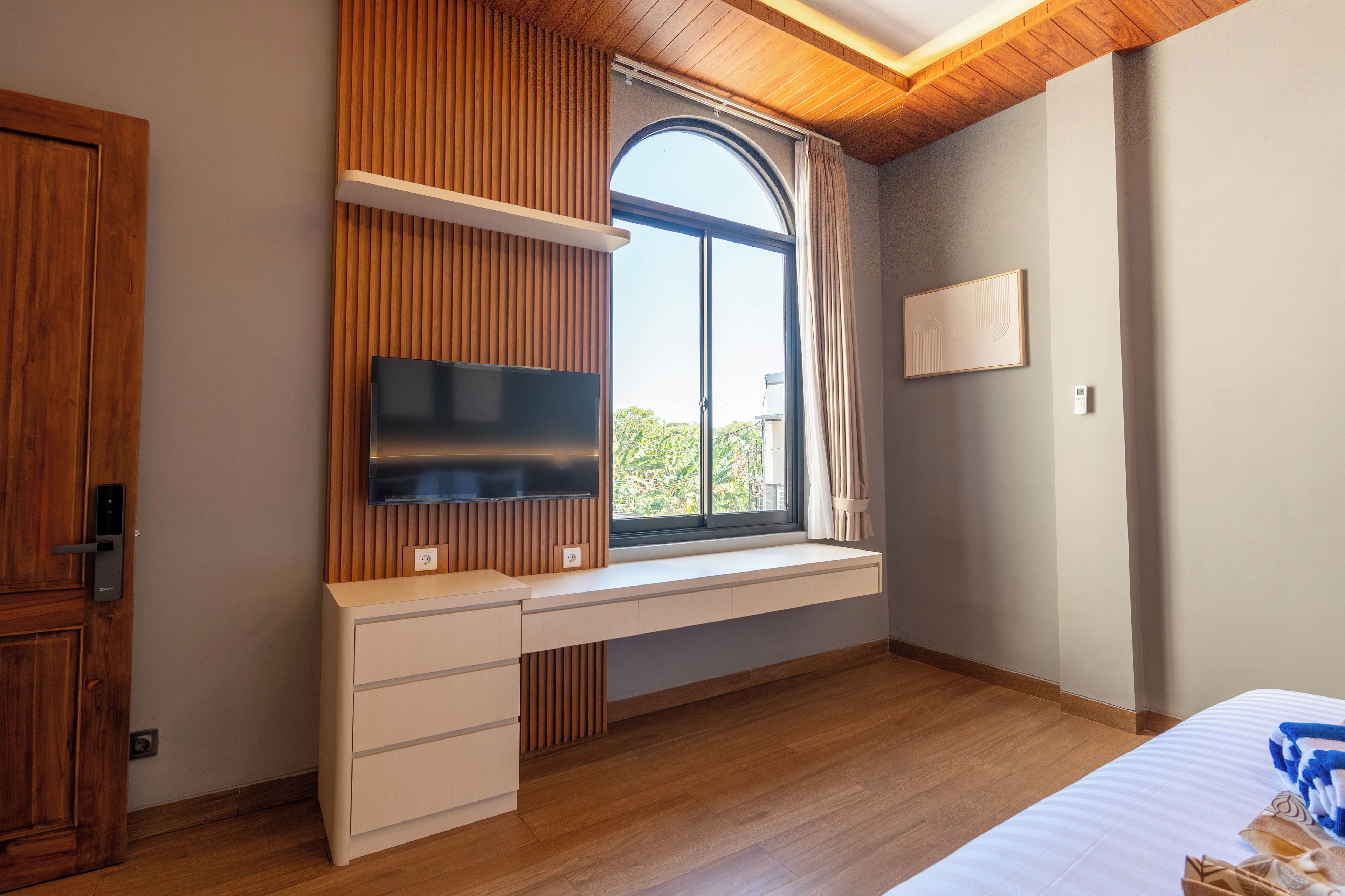A bedroom with a built-in desk, a television, a window with curtains, and a bed with a patterned blanket.