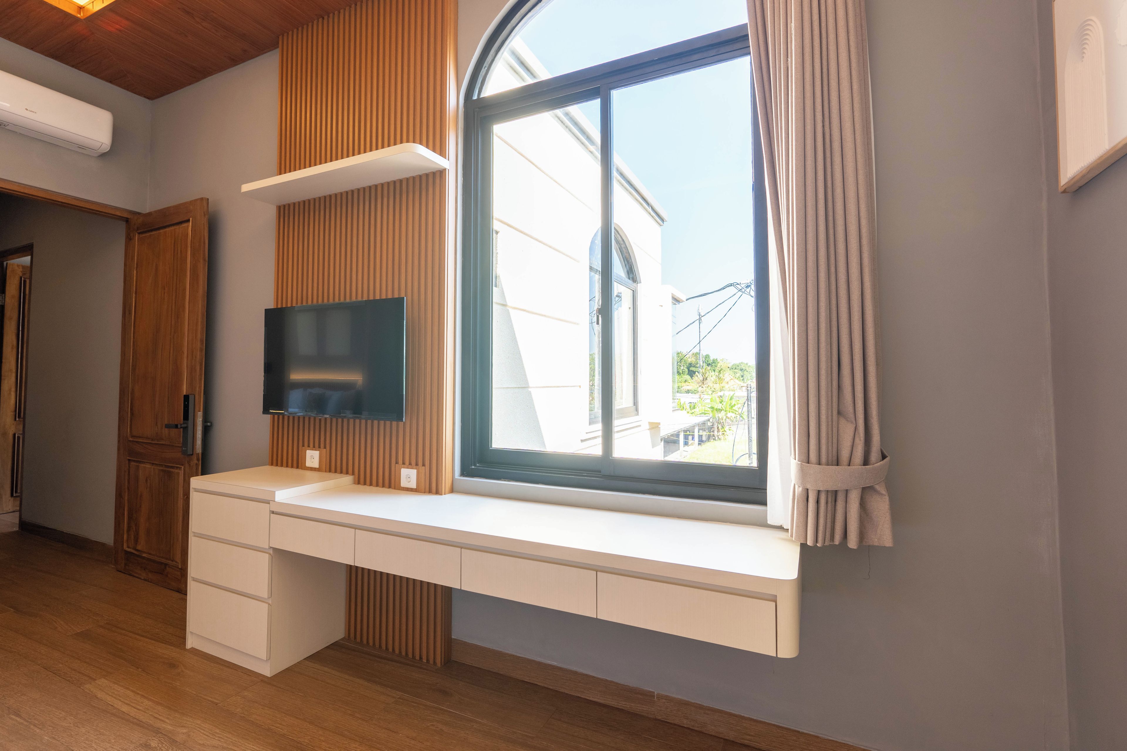 A modern room with a built-in desk, a flat-screen TV, and a large window with curtains.