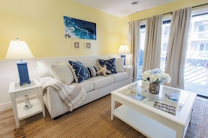 A room with a white sofa, two side tables, a glass coffee table, and a large window with curtains. - Cozy Couples Getaway! Steps to the Beach & Ocean Annies! (Myrtle Beach)