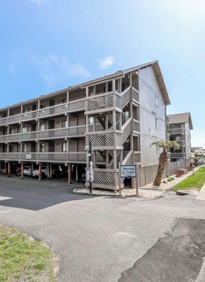 A multi-story hotel with a balcony on the second floor, a parking area in front, and a sign indicating the location. - Cozy Couples Getaway! Steps to the Beach & Ocean Annies! (Myrtle Beach)