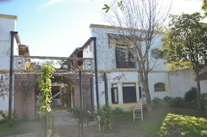 A courtyard with a pathway leading to a building entrance, surrounded by greenery and a tree. - Posada Los Angelos (Ramallo)