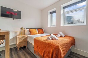 A hotel room with a bed, a TV mounted on the wall, a window, and a bedside table with a lamp. - Sutherland Apartments - Apt 41 (London)