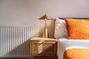 A neatly made bed with a white and orange pillow, a gold lamp on a wooden nightstand, and a white radiator. - Sutherland Apartments - Apt 41 (London)