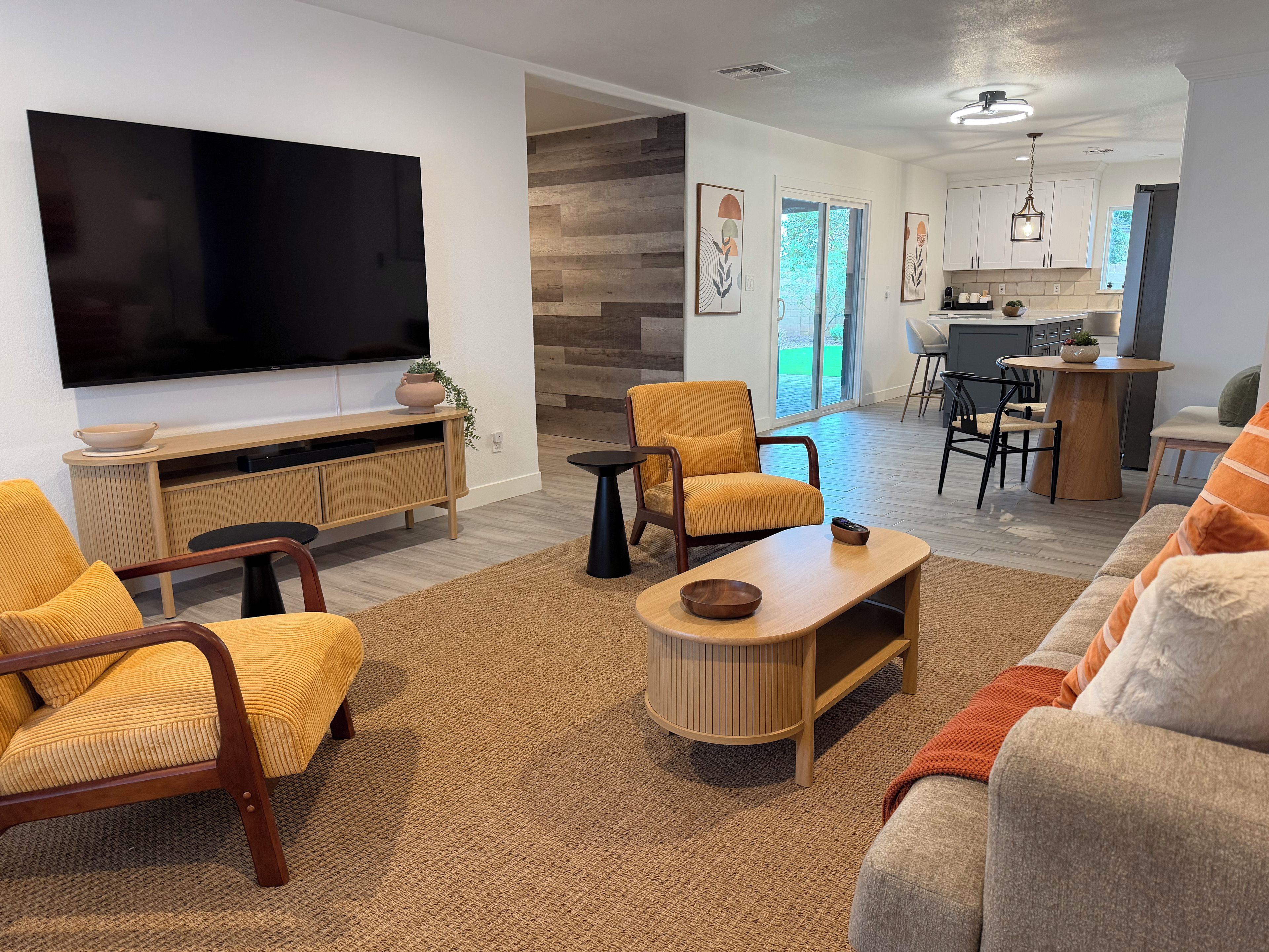 A modern living room with a flat-screen TV, a wooden entertainment unit, a sofa, and a dining area with a kitchen in the background.