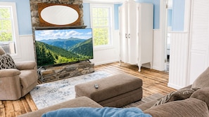 A living room with a stone fireplace, a flat-screen TV, and a large window with a mountain view. - Union Bay Cottages - The Prefix (Trenton)