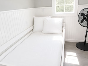 A single bed with white bedding in a room with white paneled walls and a window. - Union Bay Cottages - The Prefix (Trenton)