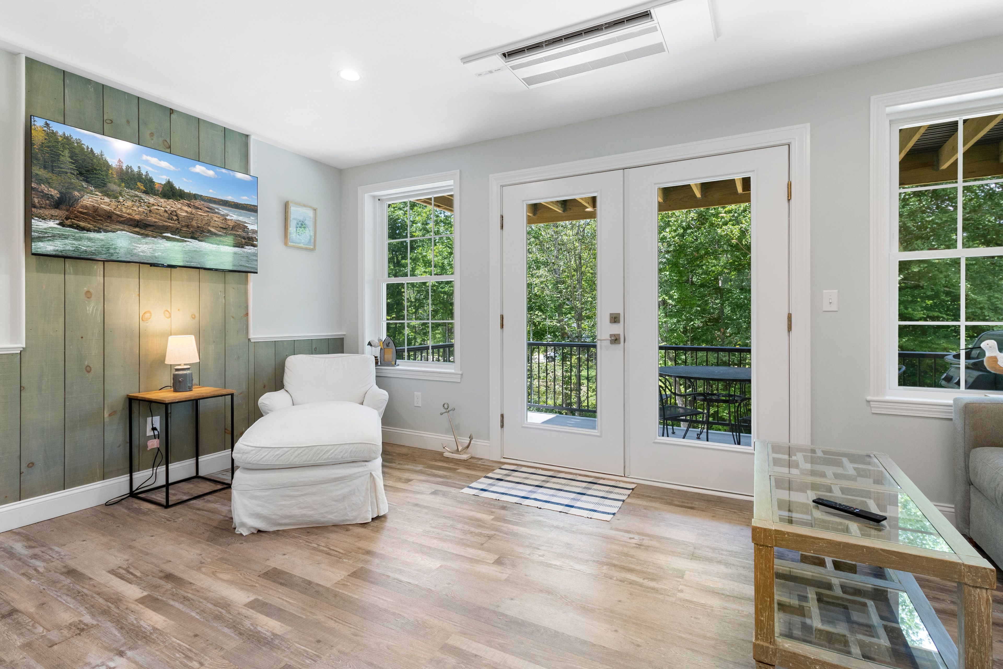 A room with a bed, a desk, a chair, a painting, and large windows with a view of trees.