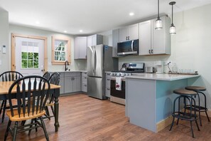 A modern kitchen with stainless steel appliances, a dining area with wooden chairs, and a view of greenery through the windows. - Blackberry Woods A Peaceful Maine Retreat in Hope (Hope)
