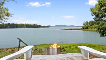 A lakeside view with a fire pit and two white chairs.